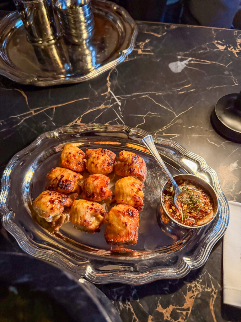 Grilled morsels on a silver platter with a dish of dressing alongside, on dark marble