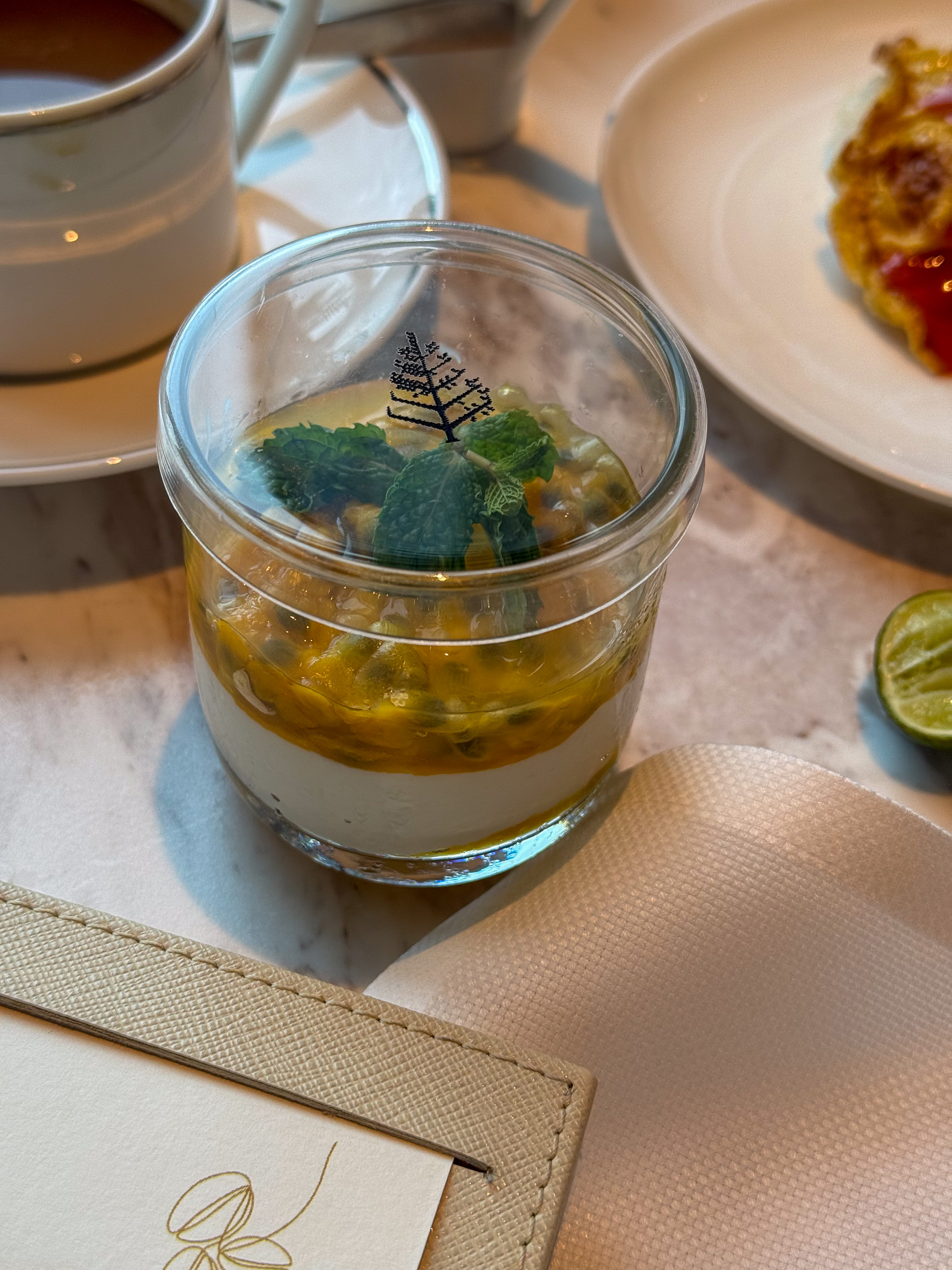 A layered dessert in a glass jar, coffee and a plate behind it