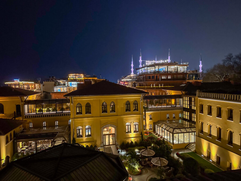 The ochre hotel lit at night from a rooftop, its glazed courtyard pavilion below and the floodlit minarets of the Blue Mosque on the ridge behind