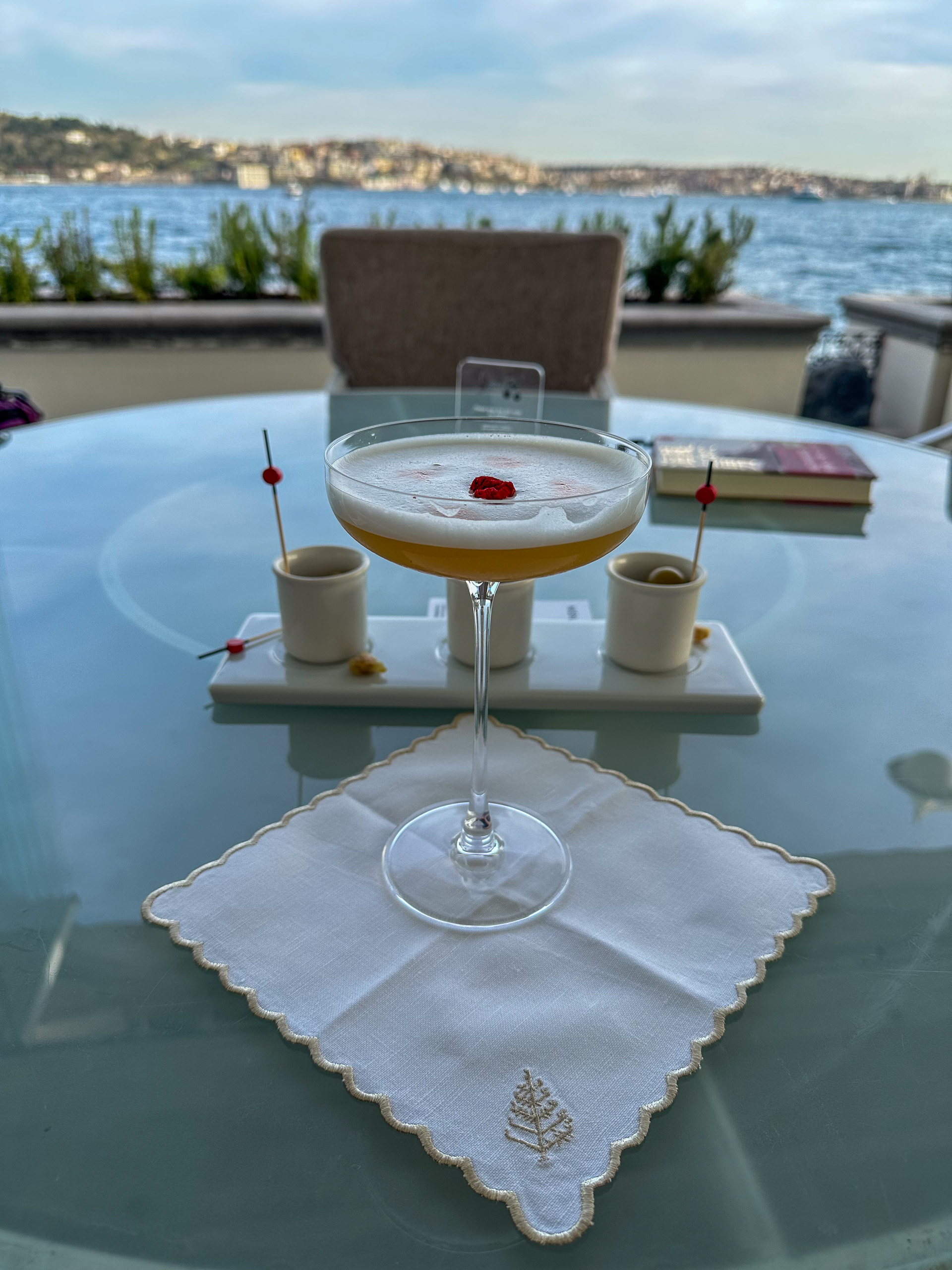 A cocktail and its accompaniments on a tray, on a glass table looking out to the water