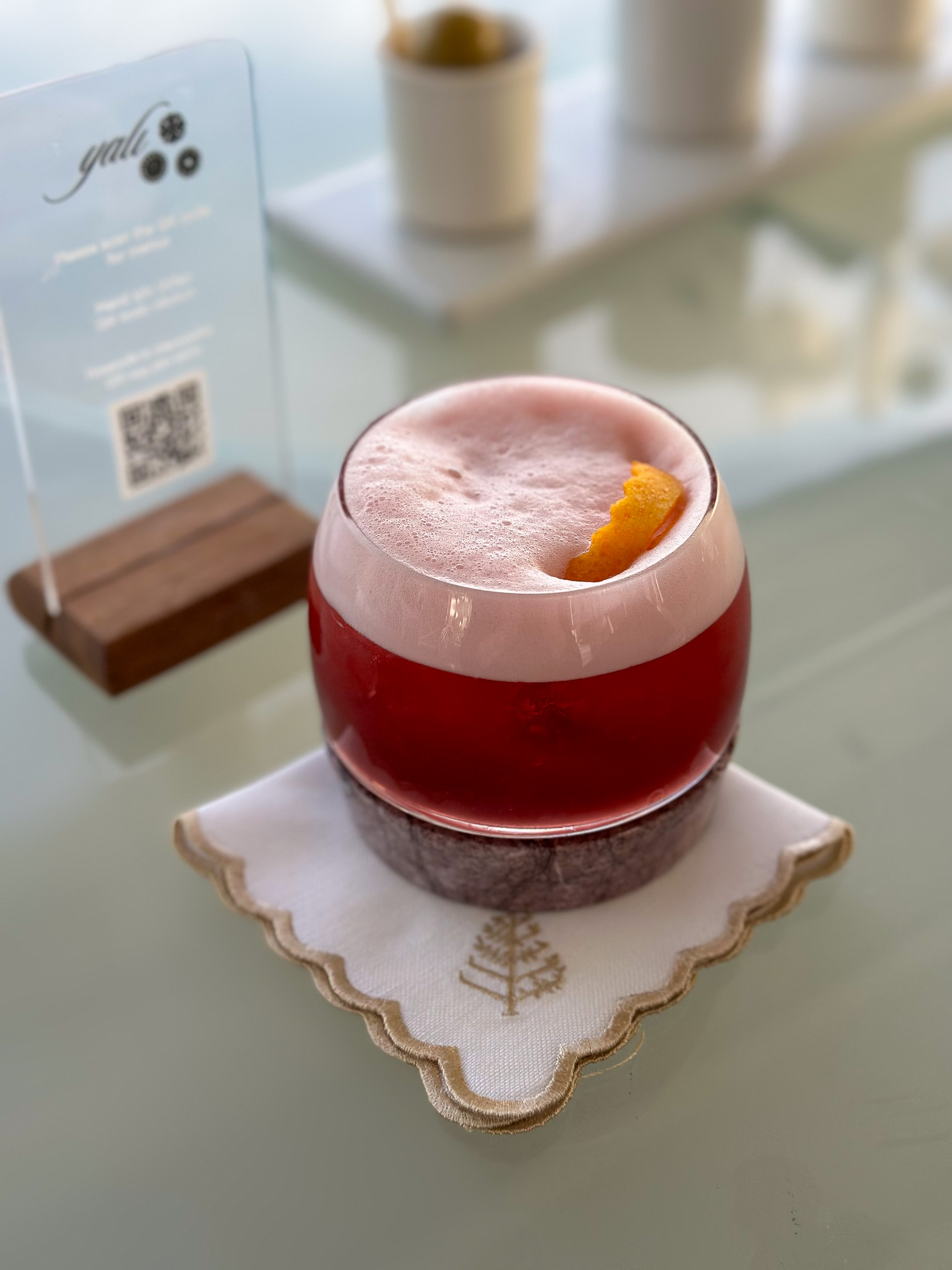A foamed pink cocktail with a citrus garnish on marble, the card standing behind it