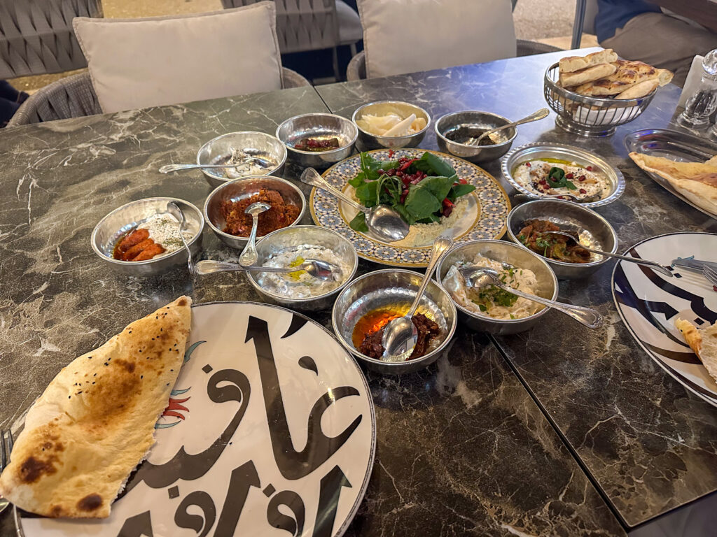 Eleven hammered steel bowls of meze ringed around a tiled plate of hummus, with a folded sesame flatbread on a calligraphy-painted plate