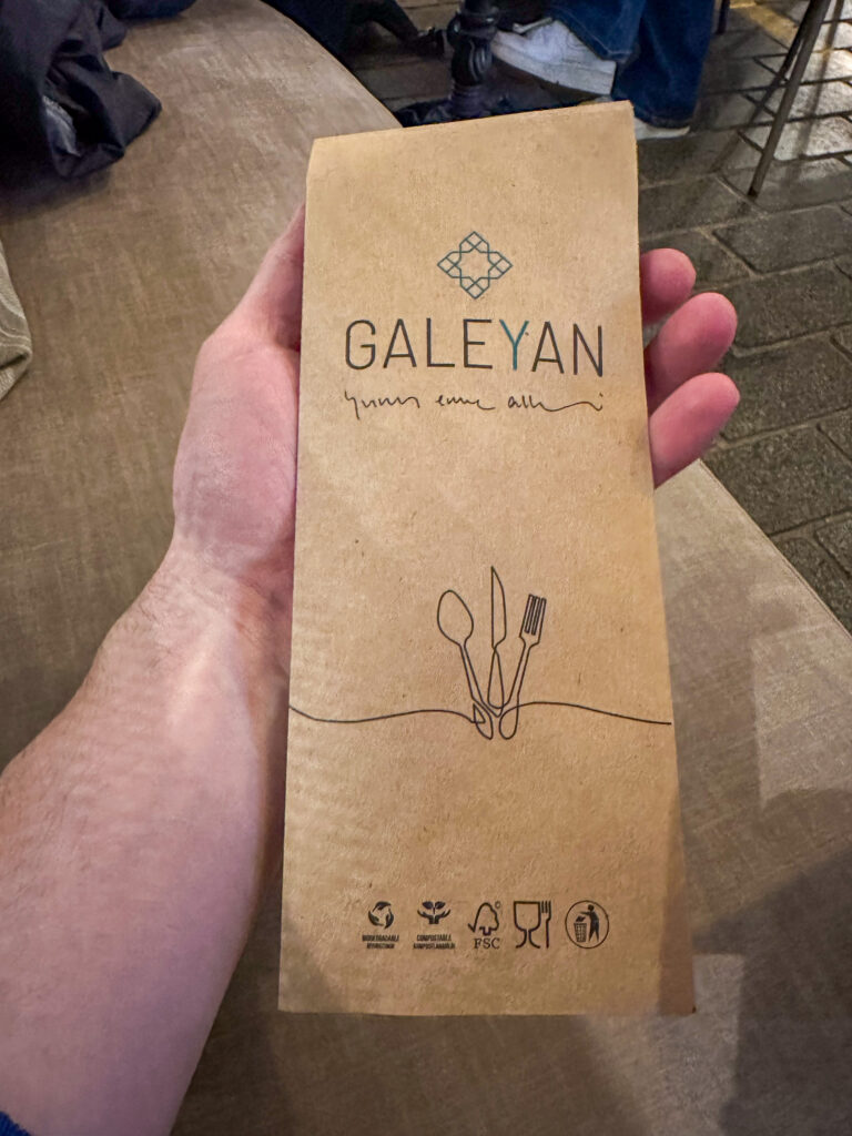 A hand holding a kraft paper cutlery sleeve printed with a tile emblem and a line drawing of a spoon, knife and fork
