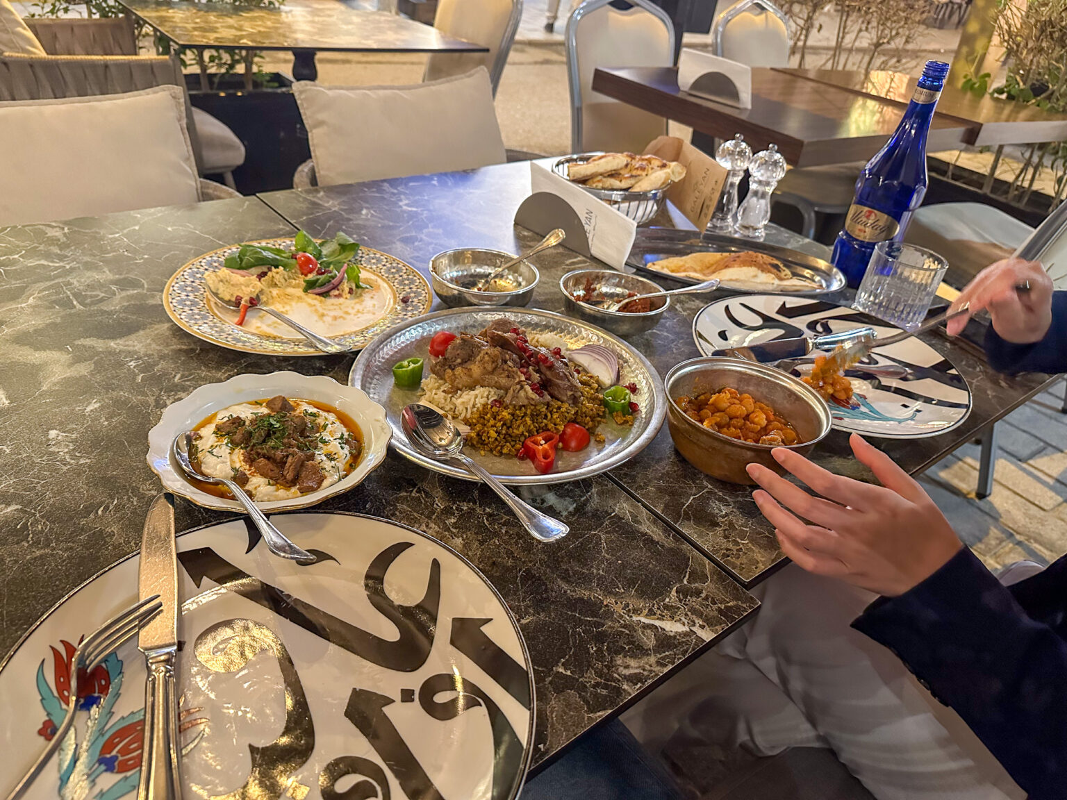 Hummus under dressed greens, a platter of grilled meat over rice and freekeh and a copper pot of chickpeas on a dark marble terrace table