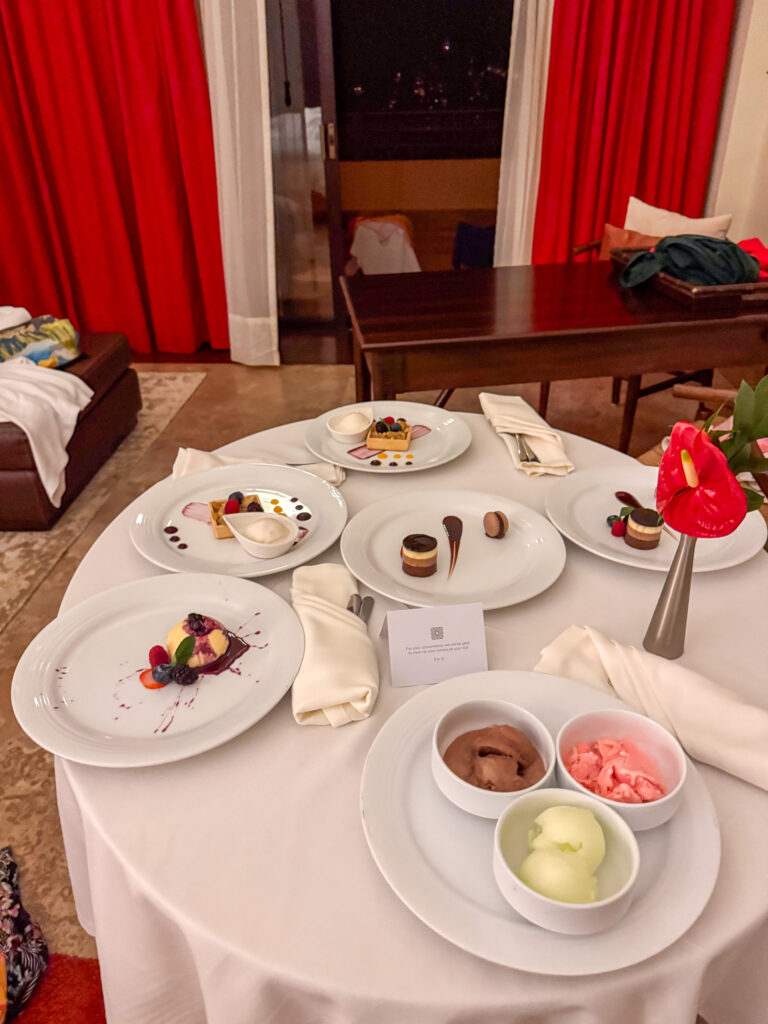 Round white-clothed table of plated desserts and sorbets beside red curtains in a darkened suite