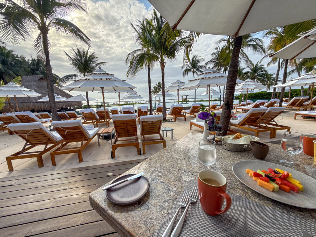 Breakfast table with coffee, juice and fruit looking out over teak loungers, parasols and palms