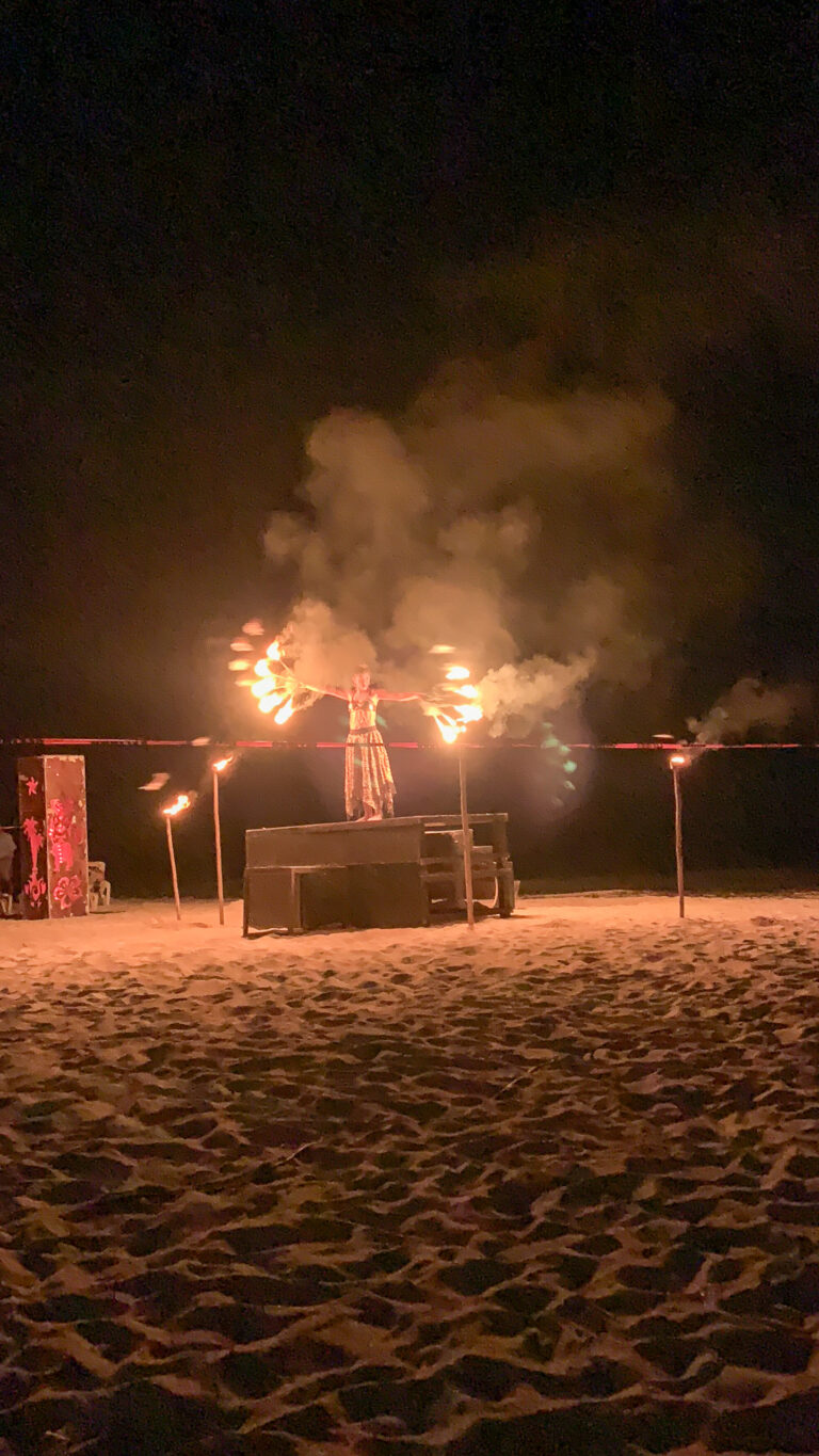 A fire performer at a distance on the dark beach, flames trailing above the sand