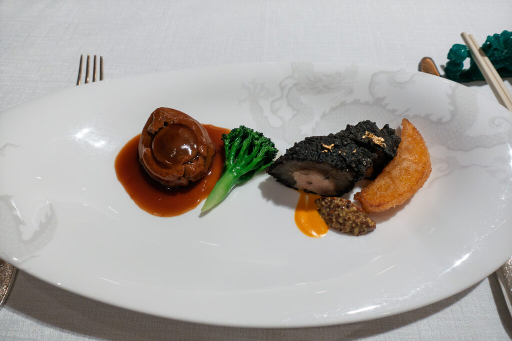 A whole braised abalone in dark sauce on a long white plate with a stem of gai lan, a gold-leafed braise, a wedge of crisp pumpkin and an orange puree