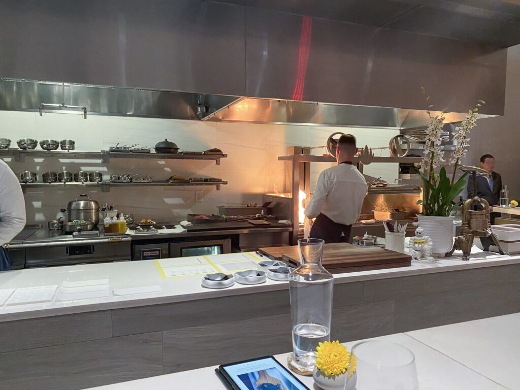 The open kitchen seen from the counter at Jônt, a cook working at the pass, a lit candle and water carafe on the white counter