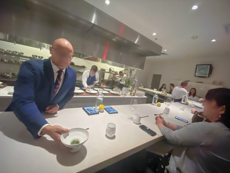 A guest in a blue suit setting down a small bowl at the counter, the kitchen working behind
