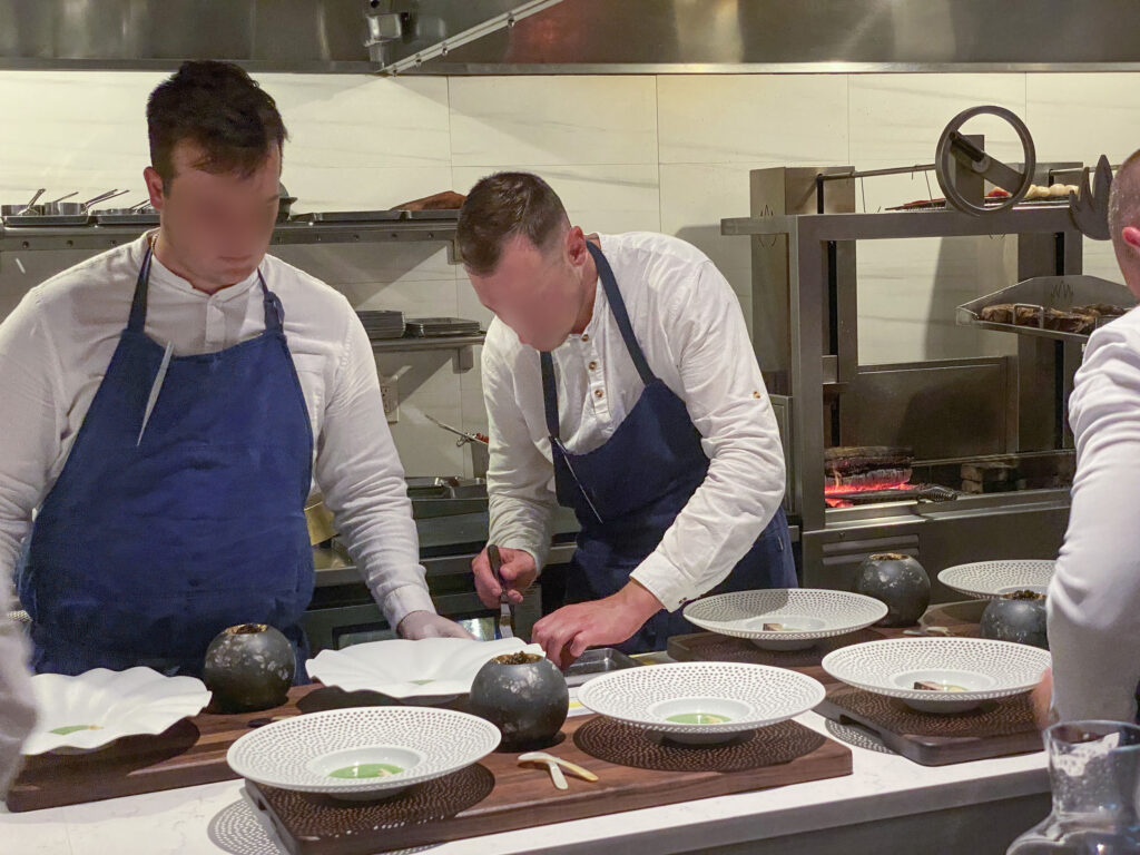 Two cooks in blue aprons plating over dark stone bowls set on a wooden board