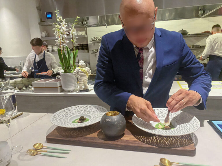 The chef in a blue suit finishing a dish at the counter, cooks working behind him