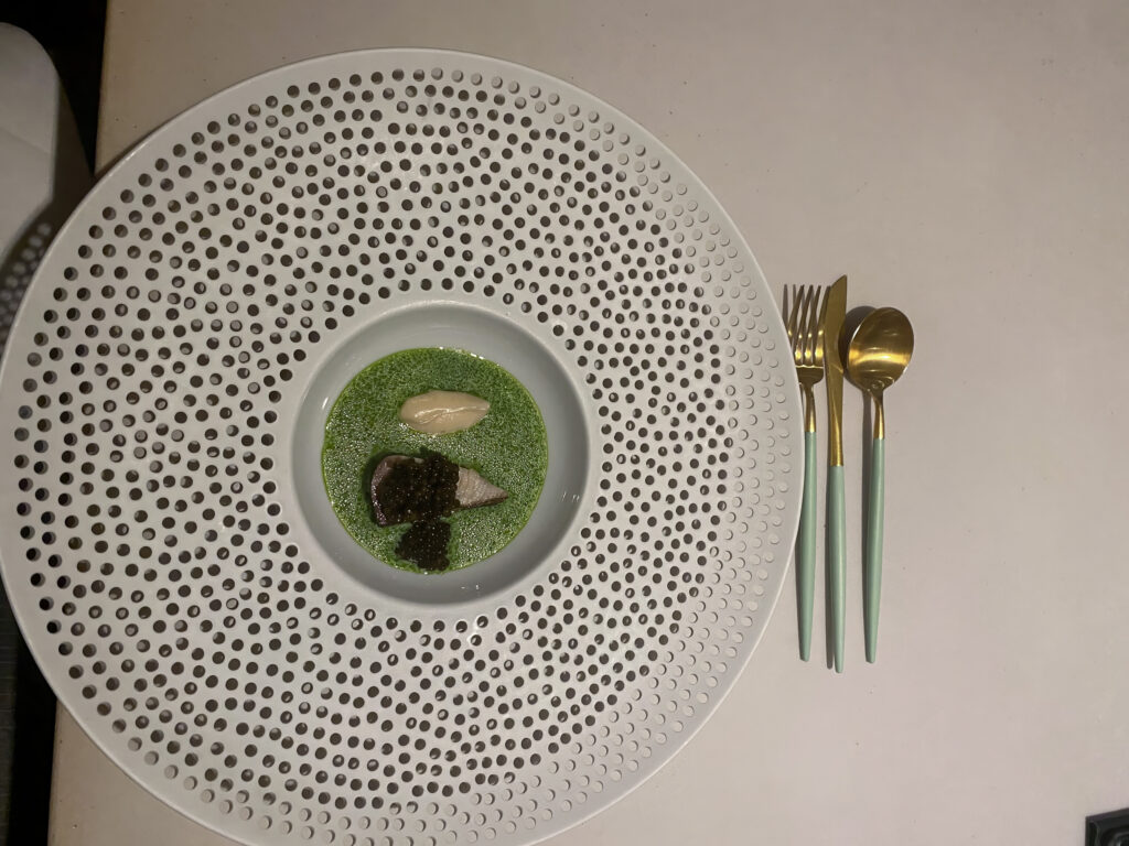 Overhead: a perforated white plate holding a green herb sauce and dark garnish, gold cutlery alongside