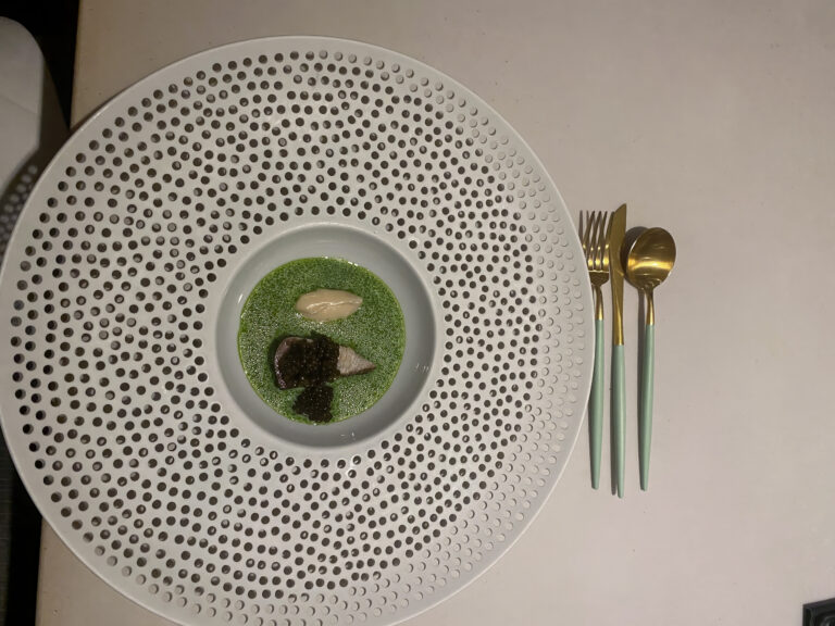 Overhead: a perforated white plate holding a green herb sauce and dark garnish, gold cutlery alongside