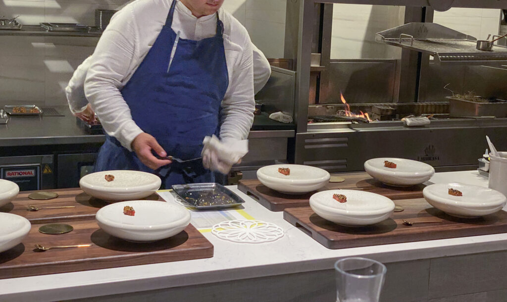 A cook in a blue apron working over a row of plates, the grill flaming behind him
