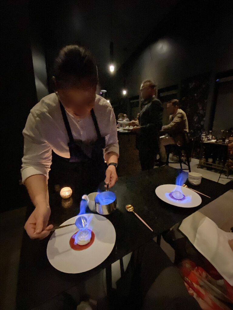A cook flaming a small copper pan beside a plate in the darkened room