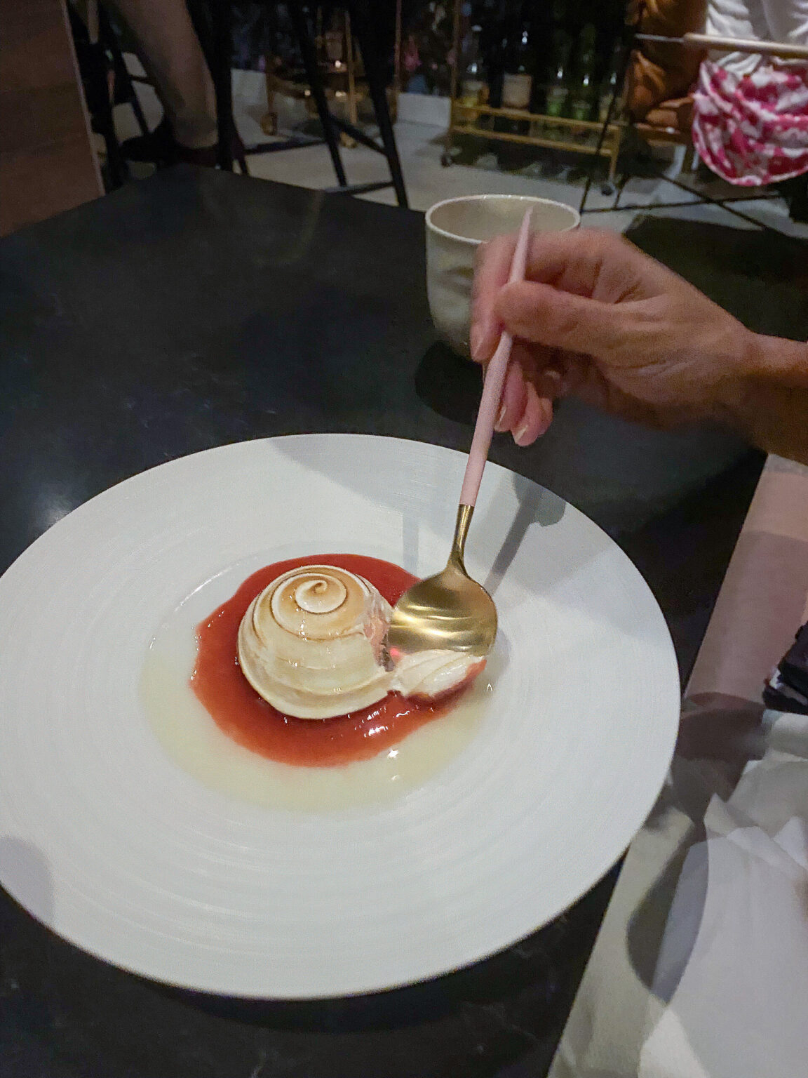 Overhead: a wide white plate with a swirl of cream set on a red fruit sauce, a gold spoon resting across it