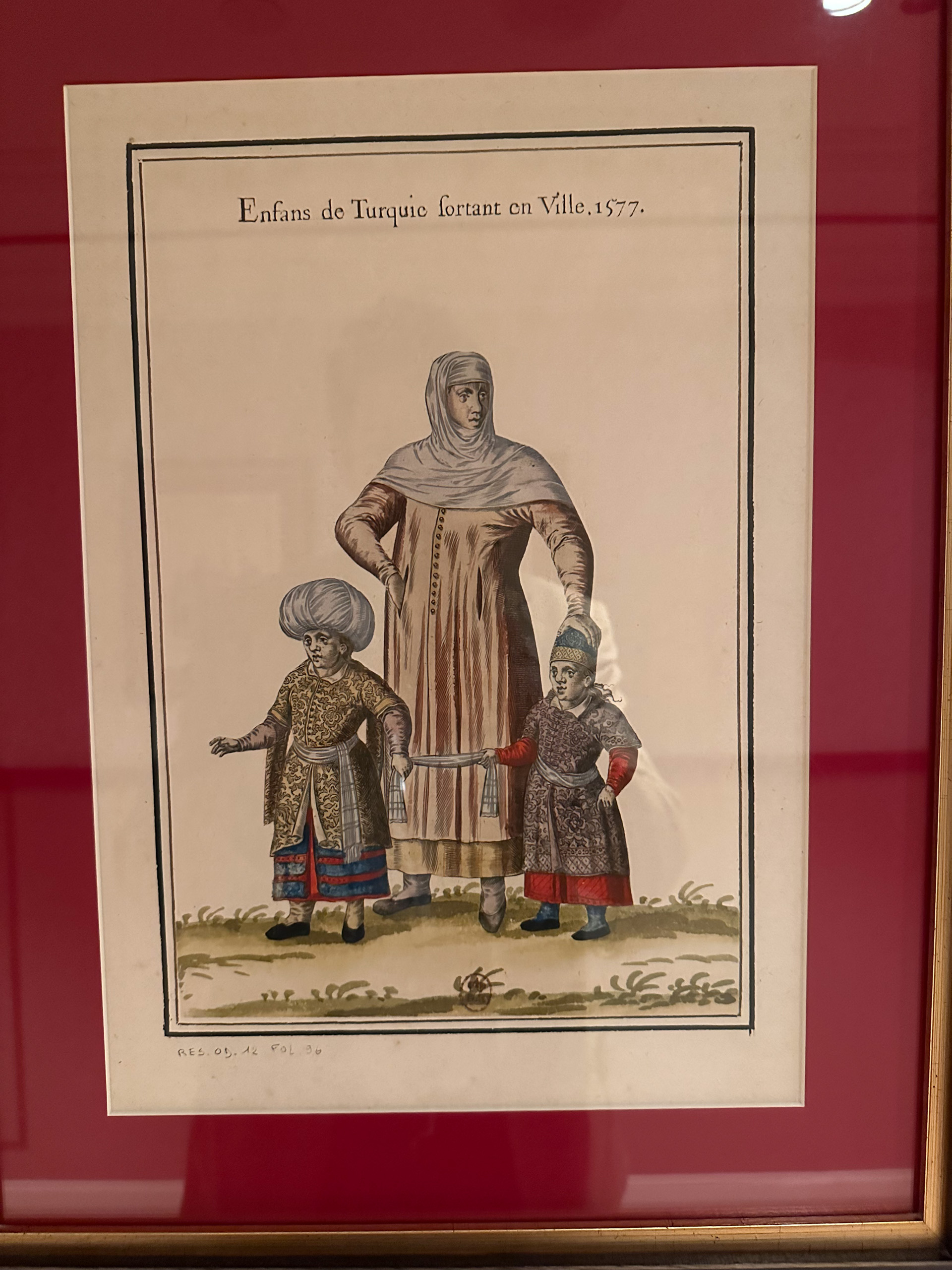 A framed hand-coloured engraving titled Enfans de Turquie sortant en Ville, dated 1577, hung on a red wall