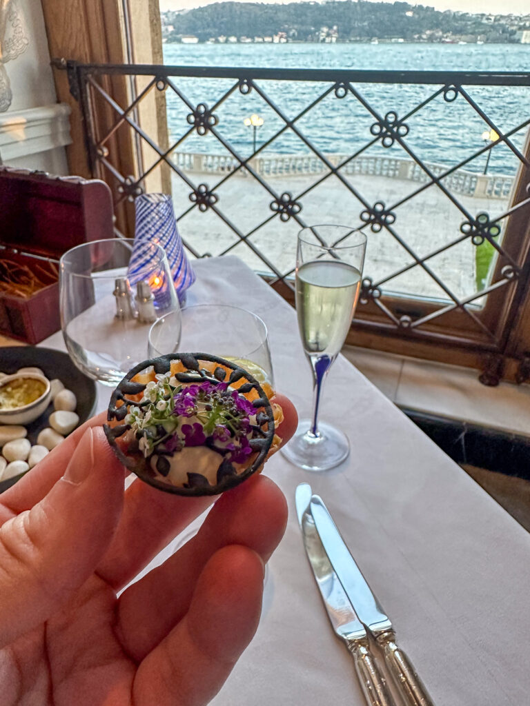 A jewelled first bite held up at the window at Tuğra, champagne and the Bosphorus behind