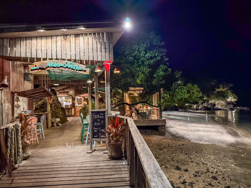 The lit timber boardwalk and thatched roof of the Walker D'Plank over-water restaurant at night, its painted sign above the bar and a table of diners on the deck beyond