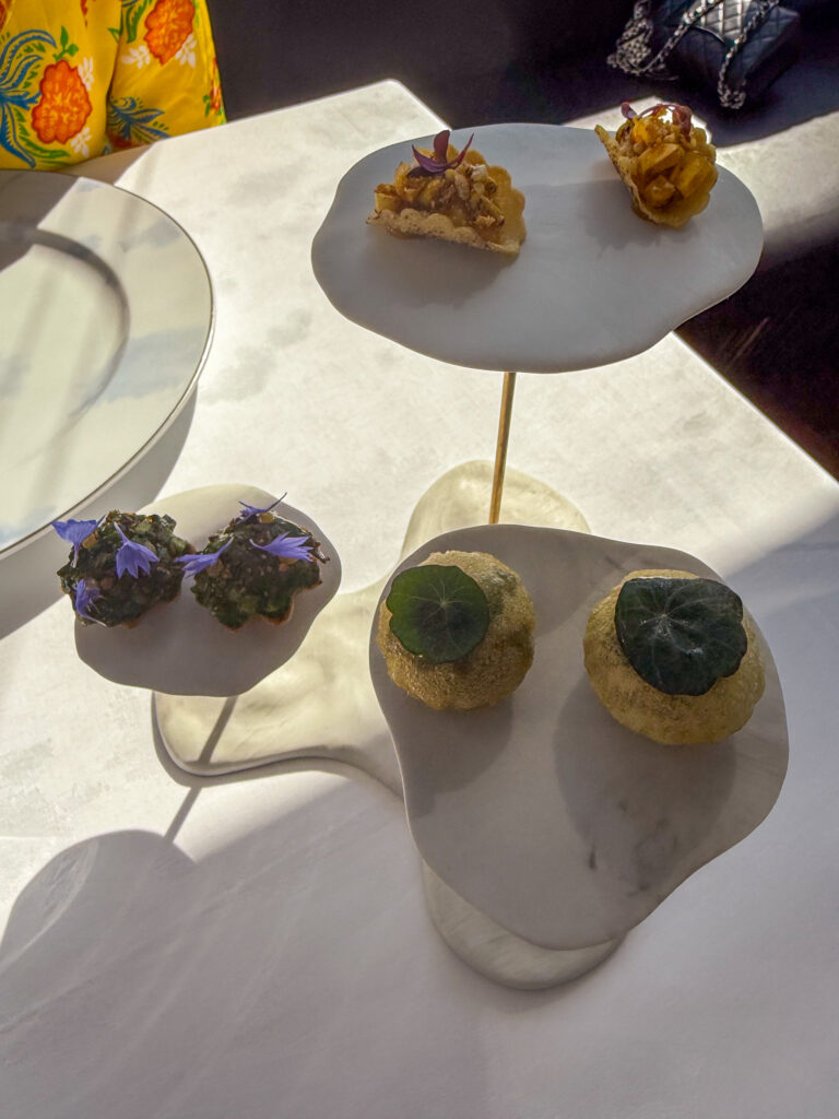 A two-tier stand of white pebble-shaped plates holding small tarts, dark canapes under blue borage flowers and pale rounds capped with nasturtium leaves