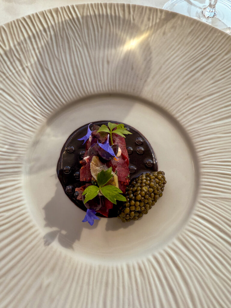 A fluted white bowl holding a dark glossy base topped with sliced pink flesh, blue borage flowers, small green leaves and a crescent of caviar