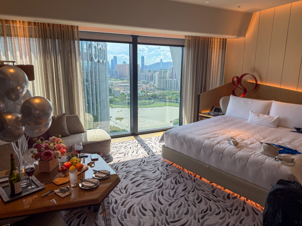 A bedroom with a pale headboard and a full-height window over Cotai and the lakes