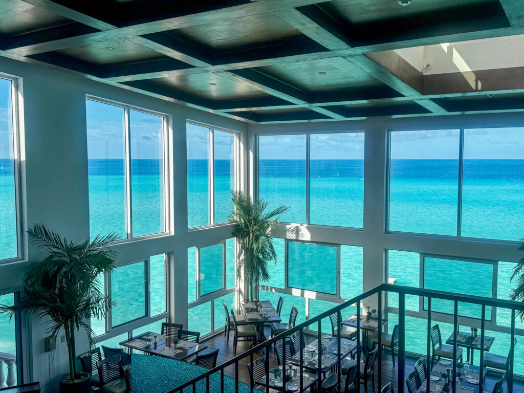The Ocean Grill dining room, double height with a coffered ceiling and floor-to-ceiling glass on two sides, tables laid and turquoise ocean filling every window
