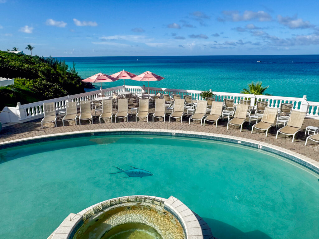 The heated pool from above, kidney-shaped and turquoise behind a white balustrade, rows of cream loungers and pink umbrellas, with a circular hot tub spilling into it