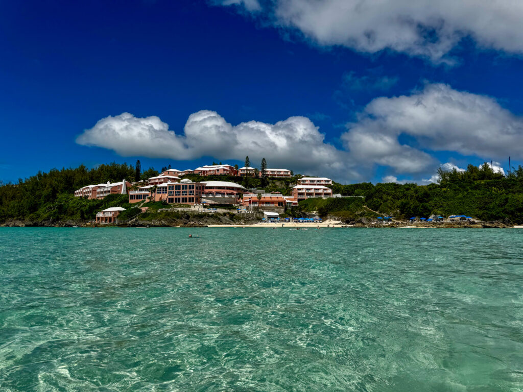 The pink and white buildings of the club stepping down a green cliff to a small beach with blue umbrellas, seen from out on the water over clear turquoise shallows