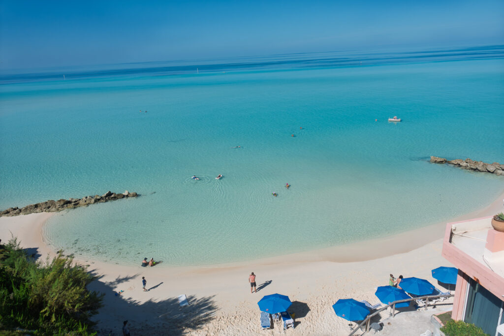 The club's beach from above, pale sand and blue umbrellas beside a rock groyne, with a vast pale turquoise shallow flat extending far out to a boat on the horizon