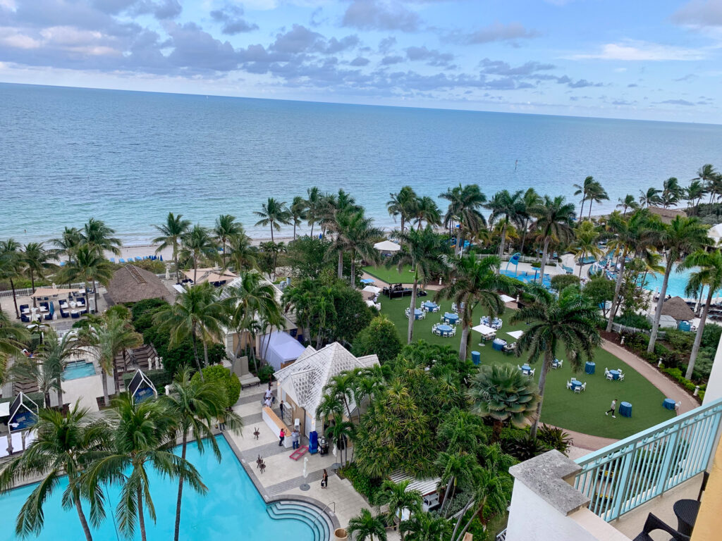 The resort from a room balcony: pools and a white pavilion below, lawns with blue loungers, palms, and the beach and Atlantic beyond under a streaked sky