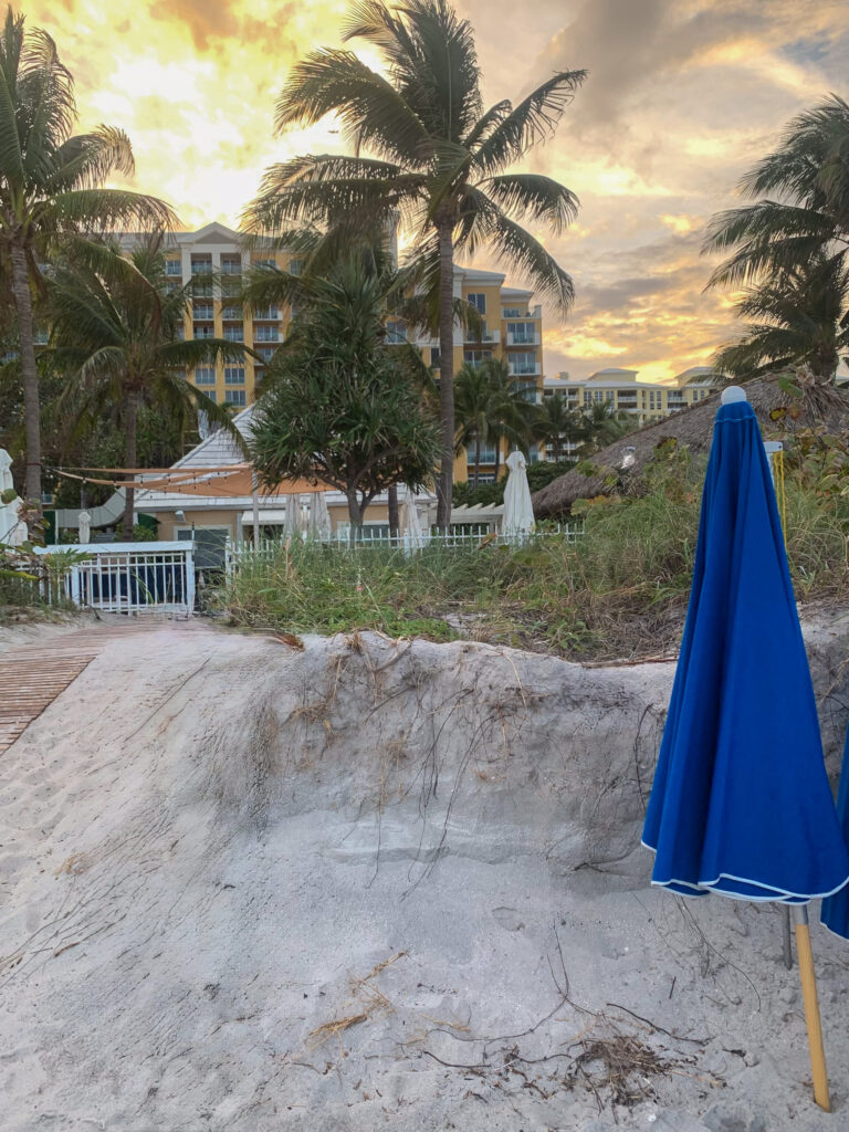A furled blue umbrella on the sand, the sun going down behind the trees