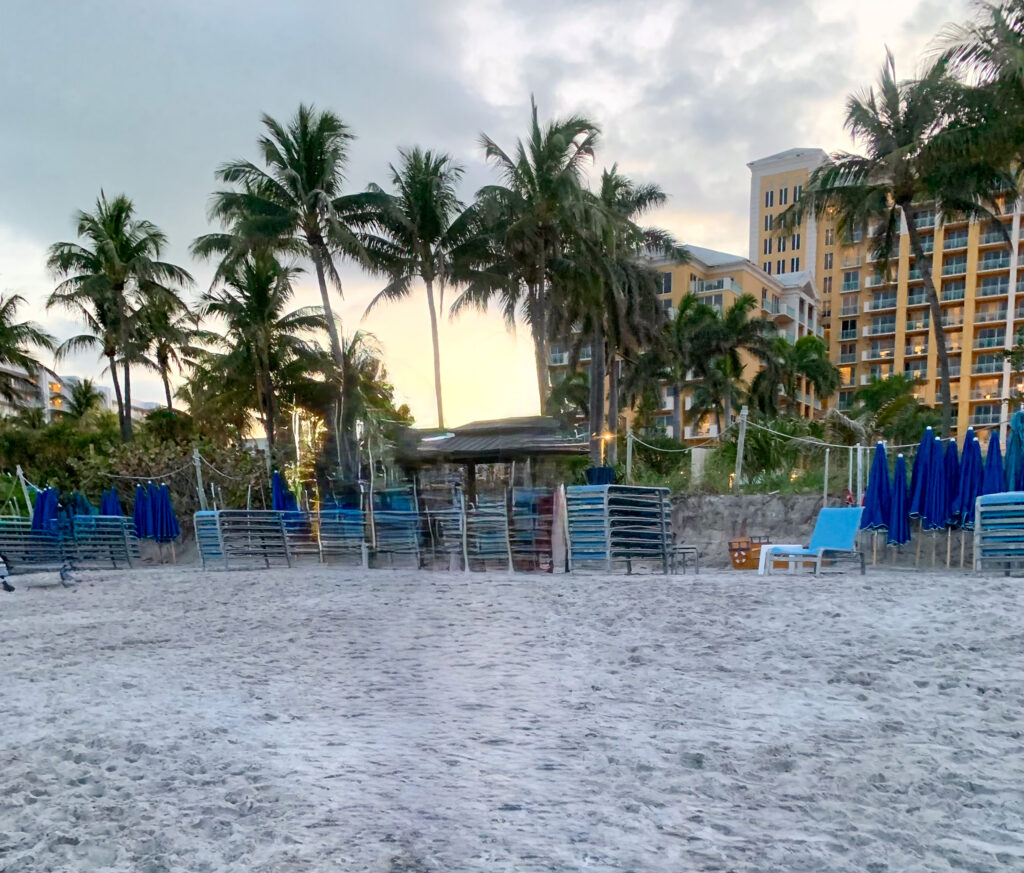 The beach at dusk, pale sand in the foreground with stacked blue loungers and folded umbrellas, palms and the resort building behind under a pale sunset