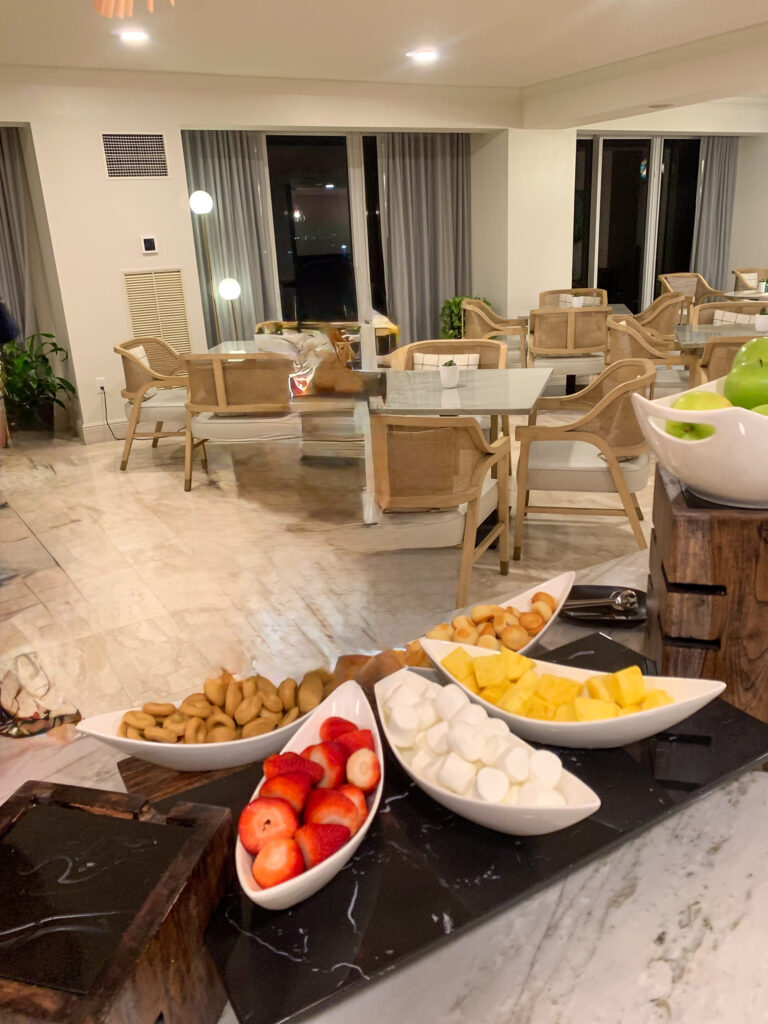 The Club Lounge counter with white dishes of strawberries, cheese, pineapple and almonds, with rattan chairs and tables in a light-filled room behind