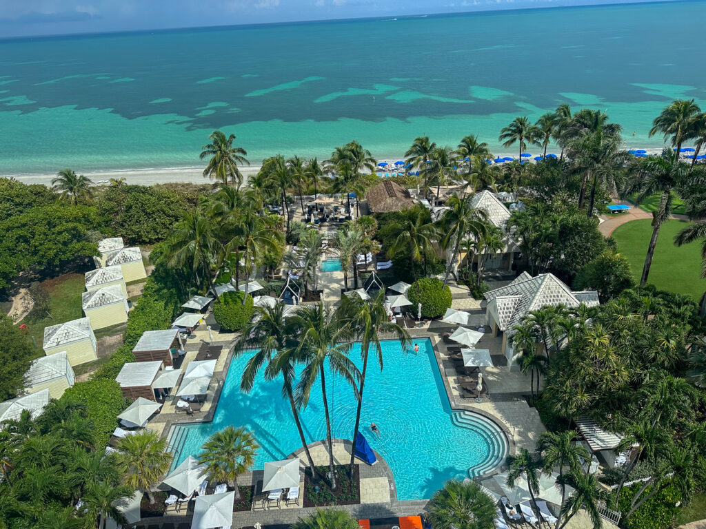 The resort seen from a high floor: a free-form turquoise pool ringed by white cabanas and palms, the beach with blue umbrellas, and shallow green Atlantic water beyond