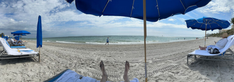 The beach framed by the edge of the umbrella and the arms of the loungers