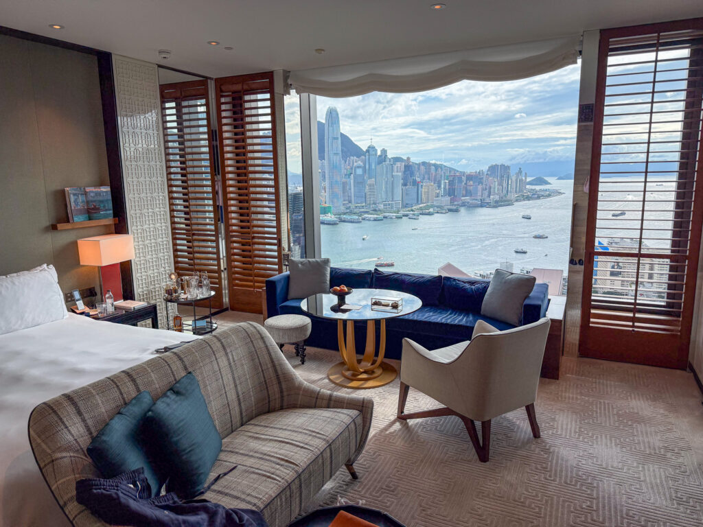 A Rosewood harbour room with a bed, a sofa and a round table by the window, plantation shutters folded back and Victoria Harbour and Hong Kong Island filling the glass