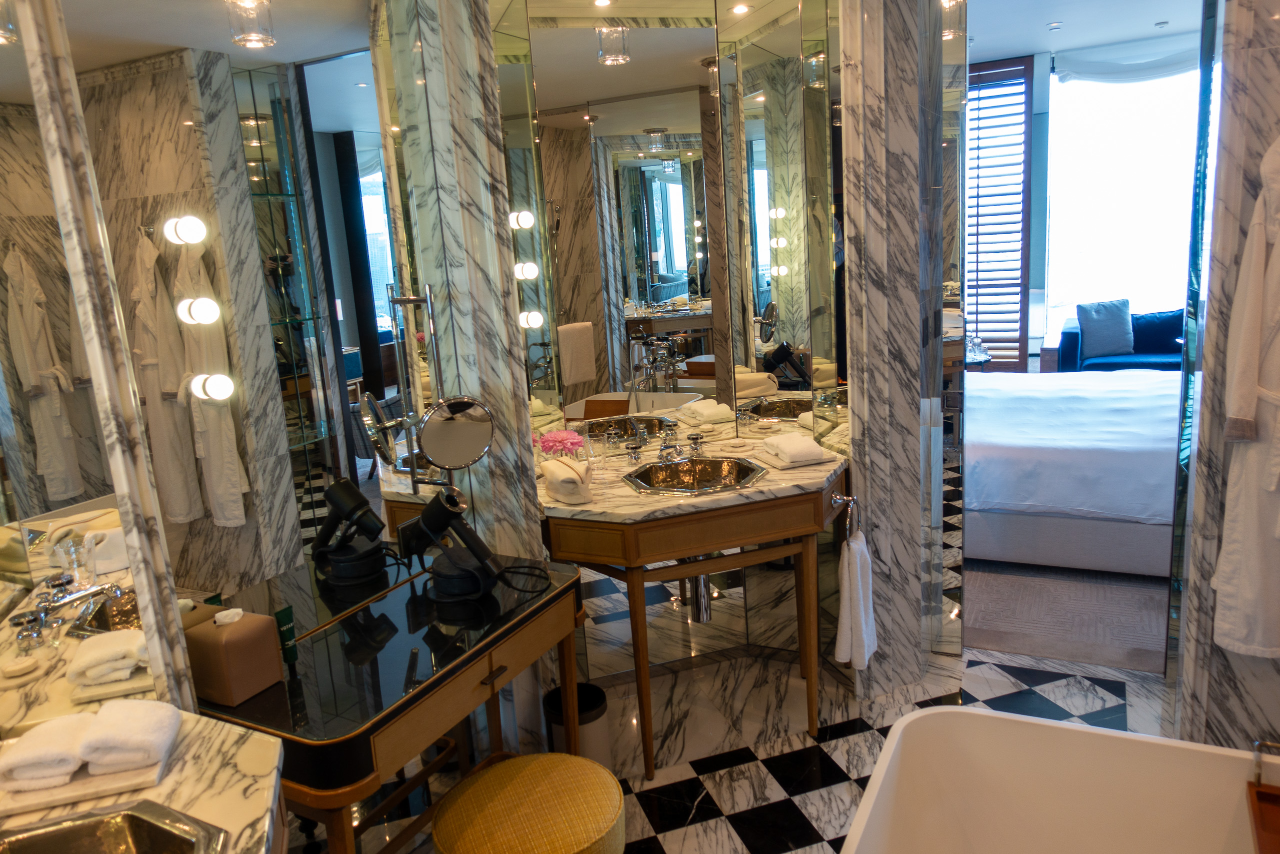 A marble bathroom with a lit vanity and a chequerboard floor, the bedroom through the door