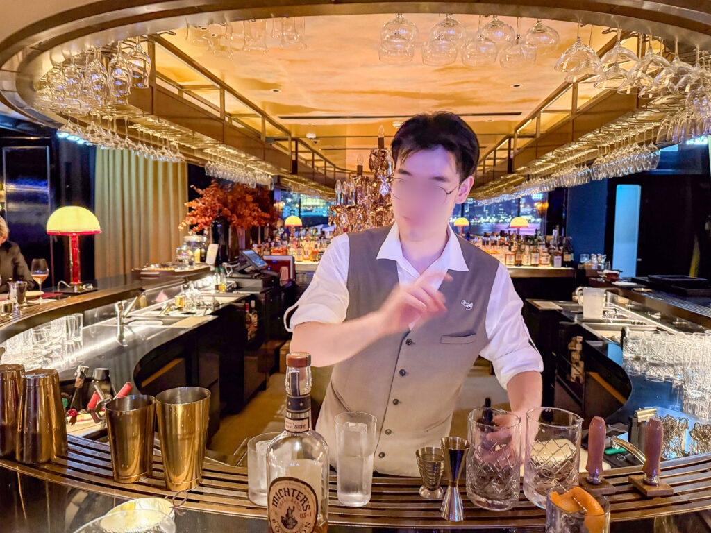 A bartender in a waistcoat mixing a drink at a low-lit bar with a gilded ceiling, bottles and shakers across the counter