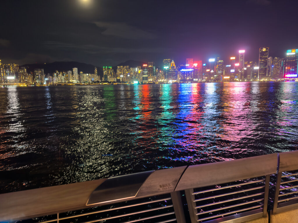 The Hong Kong Island skyline lit at night in full colour, reflected the length of Victoria Harbour, seen from a Kowloon terrace