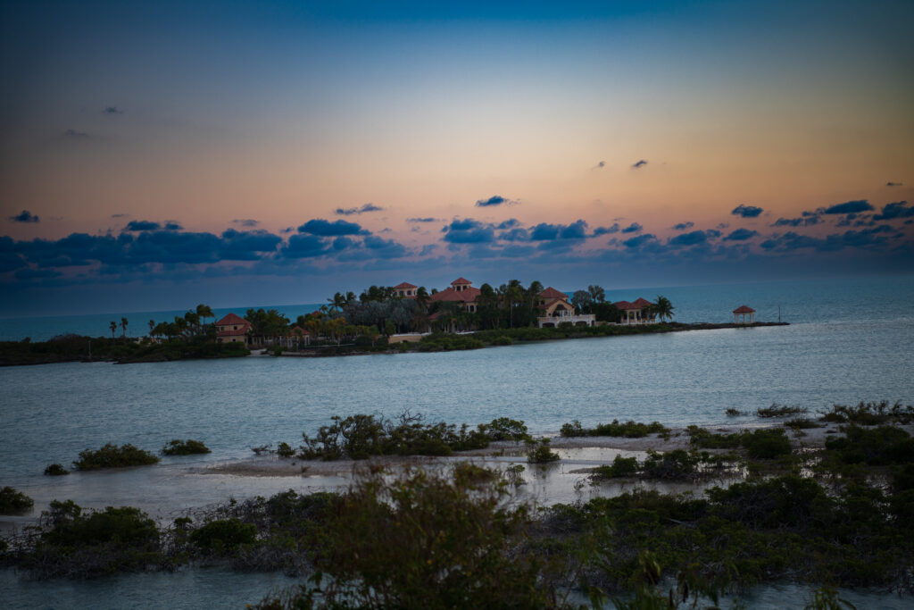 A low wooded point at dusk with its houses lit, a small pavilion at the water's edge and the sea flat to the horizon