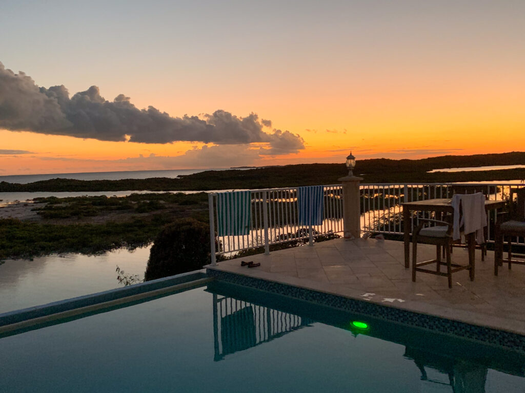 A villa's infinity pool and railed terrace at sunset, striped towels drying on the rail and a bar-height table set beside it