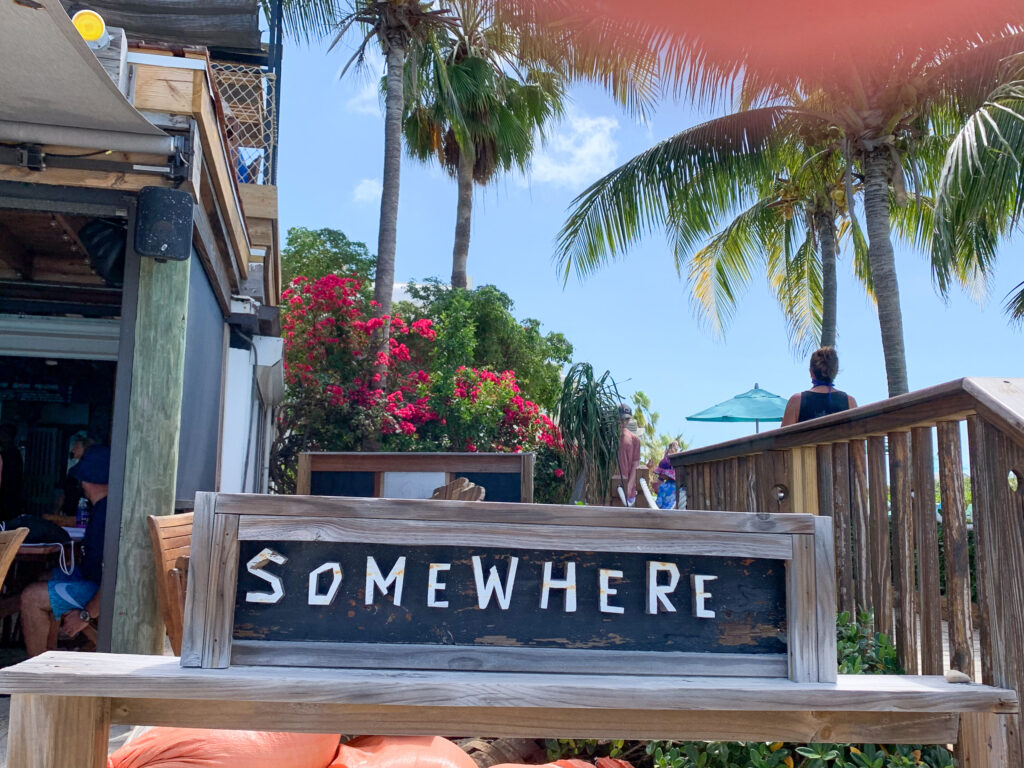 A driftwood board spelling SOMEWHERE in white shell lettering propped on a bench in front of the timber bar, with bougainvillea and palms behind