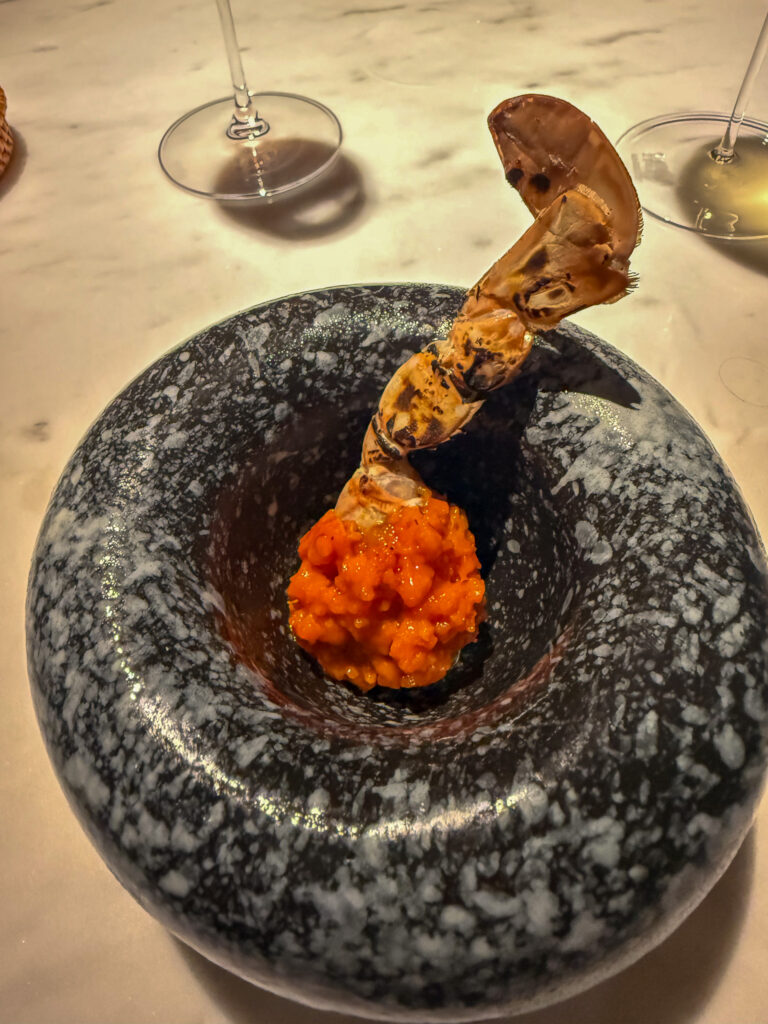 A mound of bright orange roe with a charred prawn tail arching out of it, served in a heavy speckled black stone bowl between two wine glasses