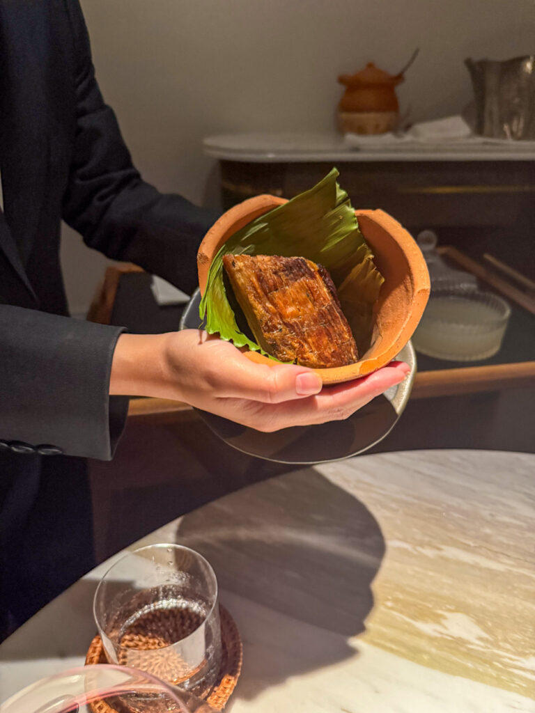 A hand holding a broken clay pot lined with banana leaf, a piece of charred grilled fish inside