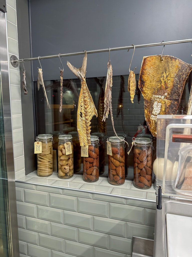 Whole fish hung to dry on a rail beside glass jars of preserved figs and dried chillies